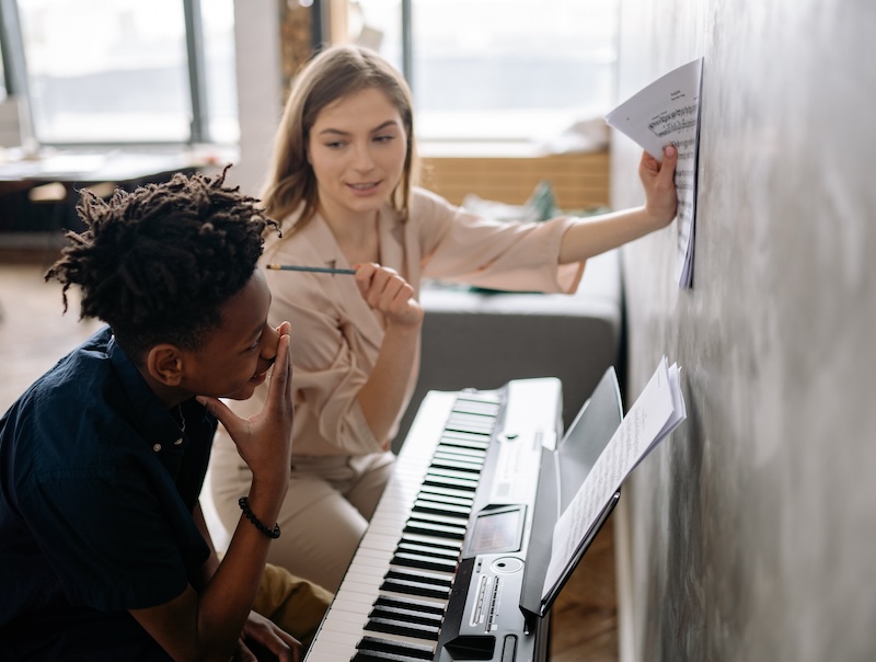 Piano-docent houdt muziekpapier tegen de muur terwijl ze naar haar piano-leerling kijkt, die geconcentreerd luistert naar wat zijn juf te zeggen heeft.
